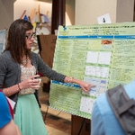 A student presenter holding a cup and pointing out something on her poster as she presents